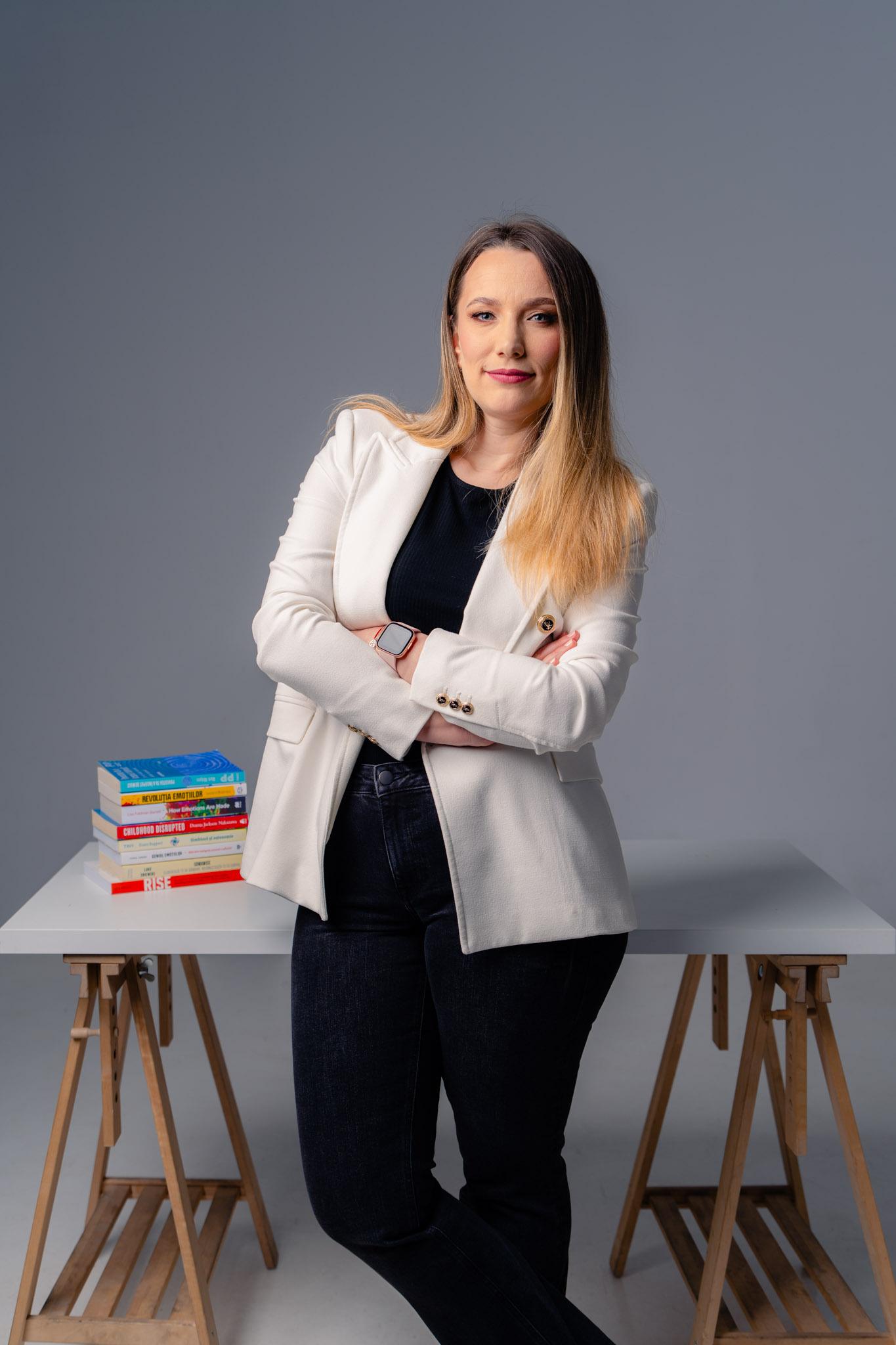 Andreea Negoiță - coach de inteligență emoțională, portret în sacou alb, fundal întunecat, cărți pe birou