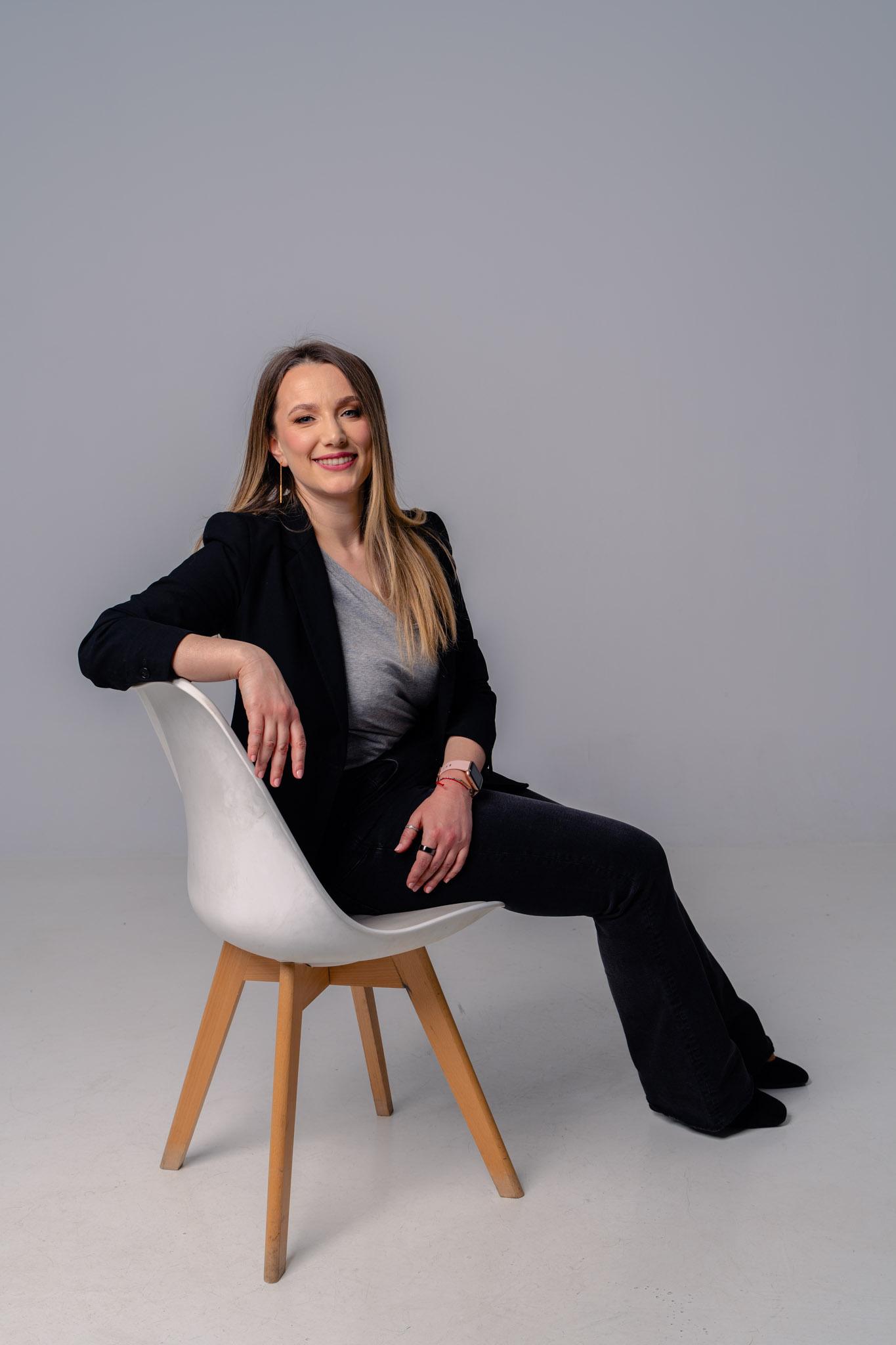 Andreea Negoiță - coach de inteligență emoțională, portret business, ținută neagră, așezată pe scaun alb, fundal gr