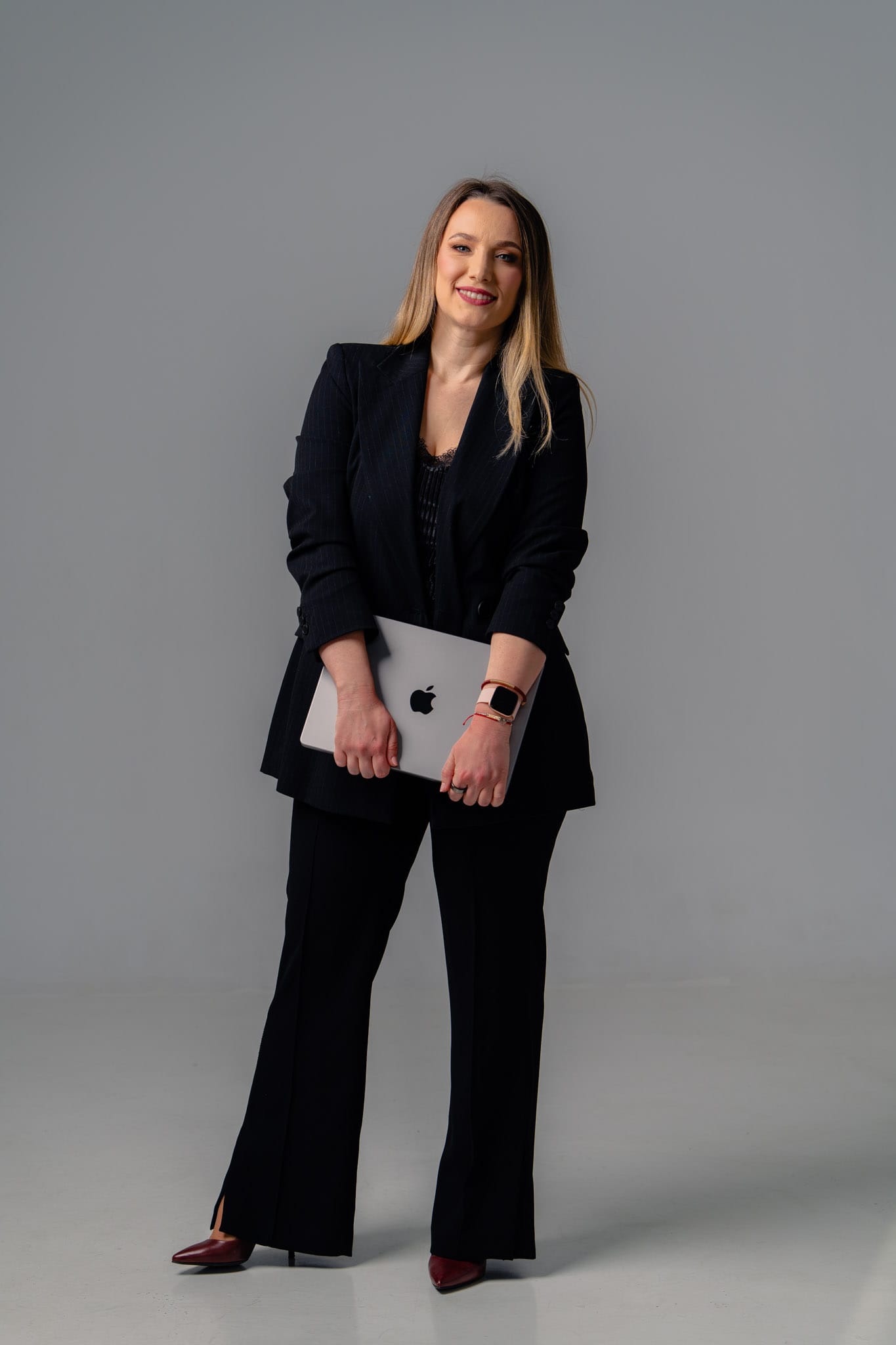 Andreea Negoiță - coach de inteligență emoțională, portret business, sacou negru, în picioare, ținând un laptop, fundal neutru